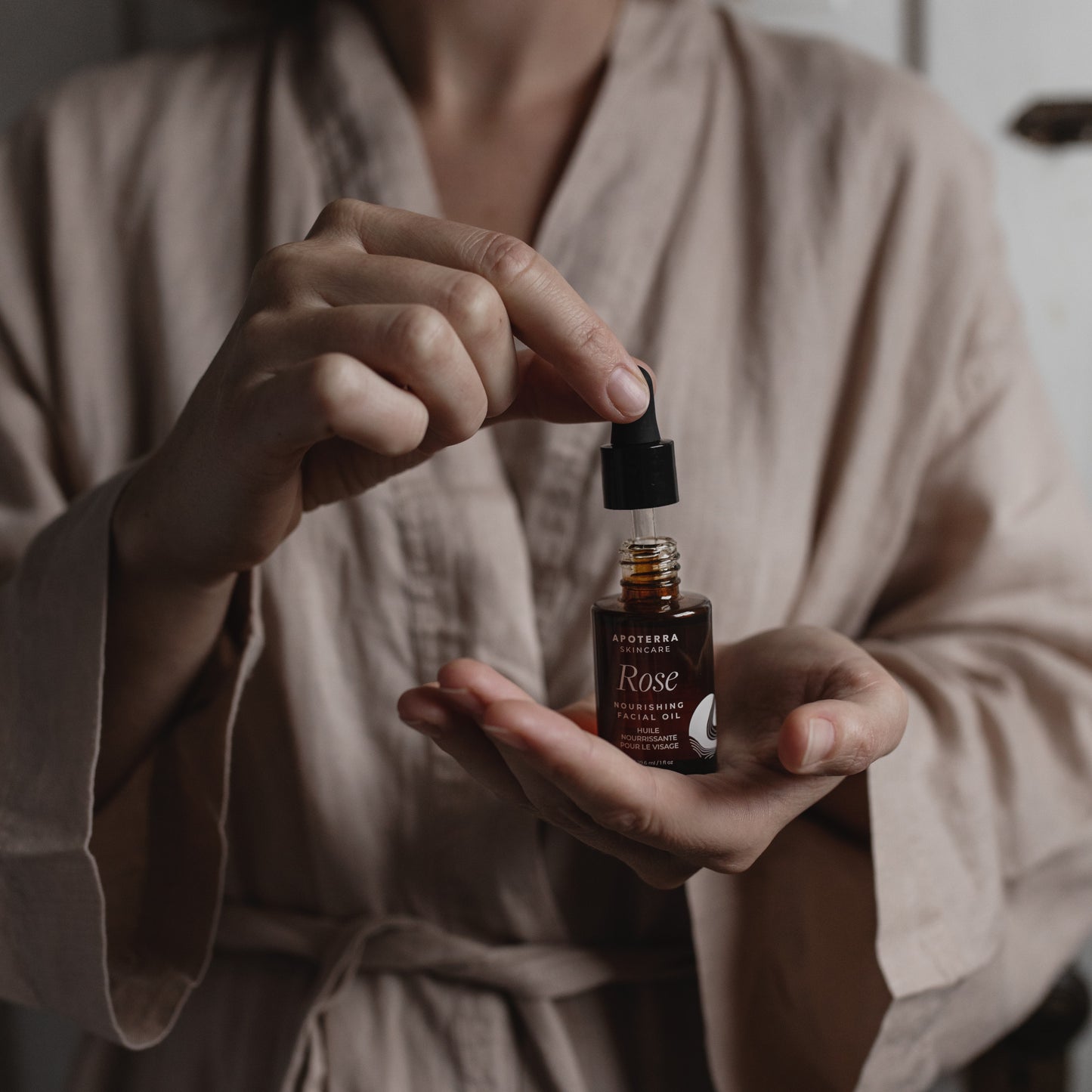 Person holding a dropper bottle of rose facial oil in a robe. APOTERRA Rose Nourishing Facial Oil
