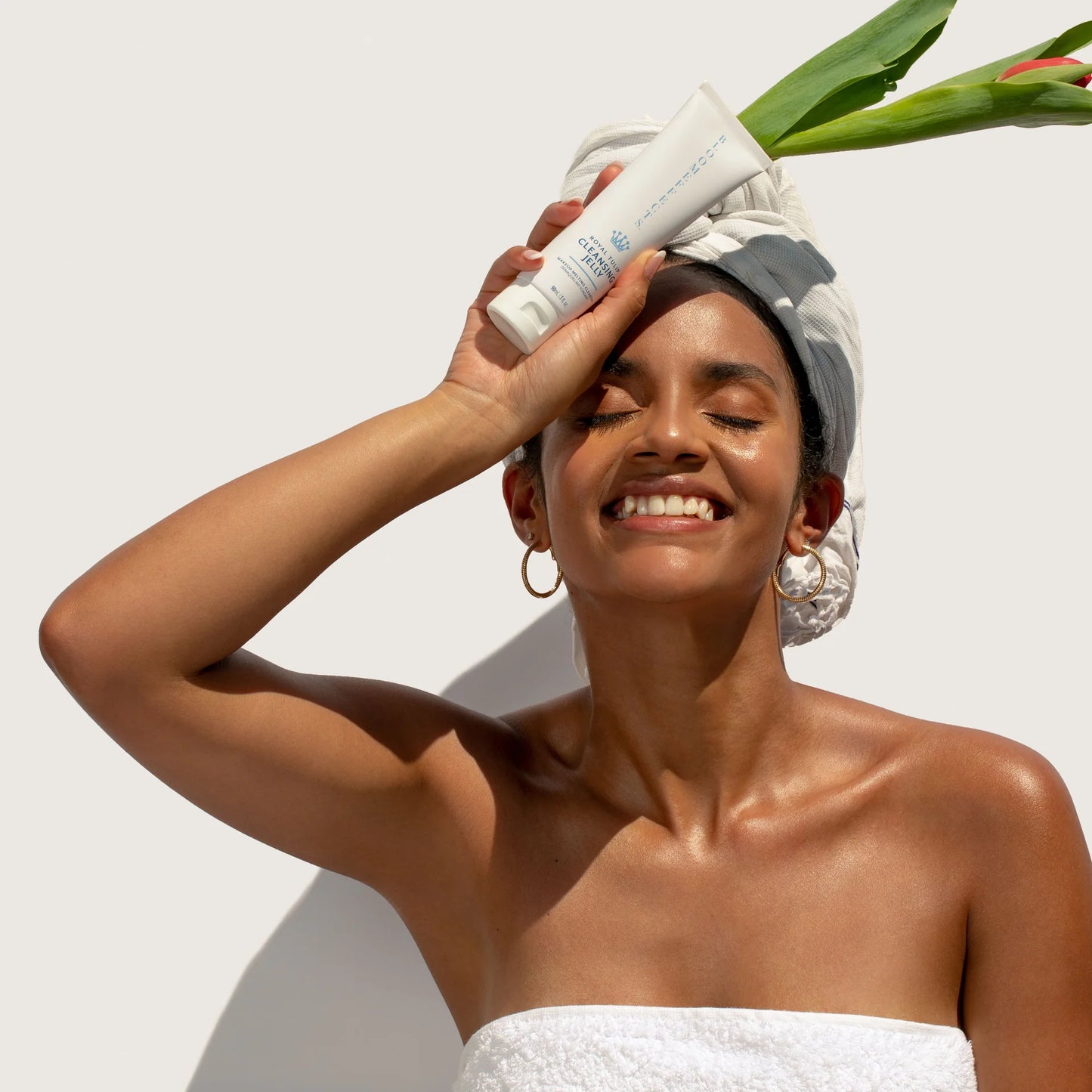 Woman with a towel wrapped around her hair holding a white tube of BLOOMEFFECTS Royal Tulip Cleansing Jelly against a white background