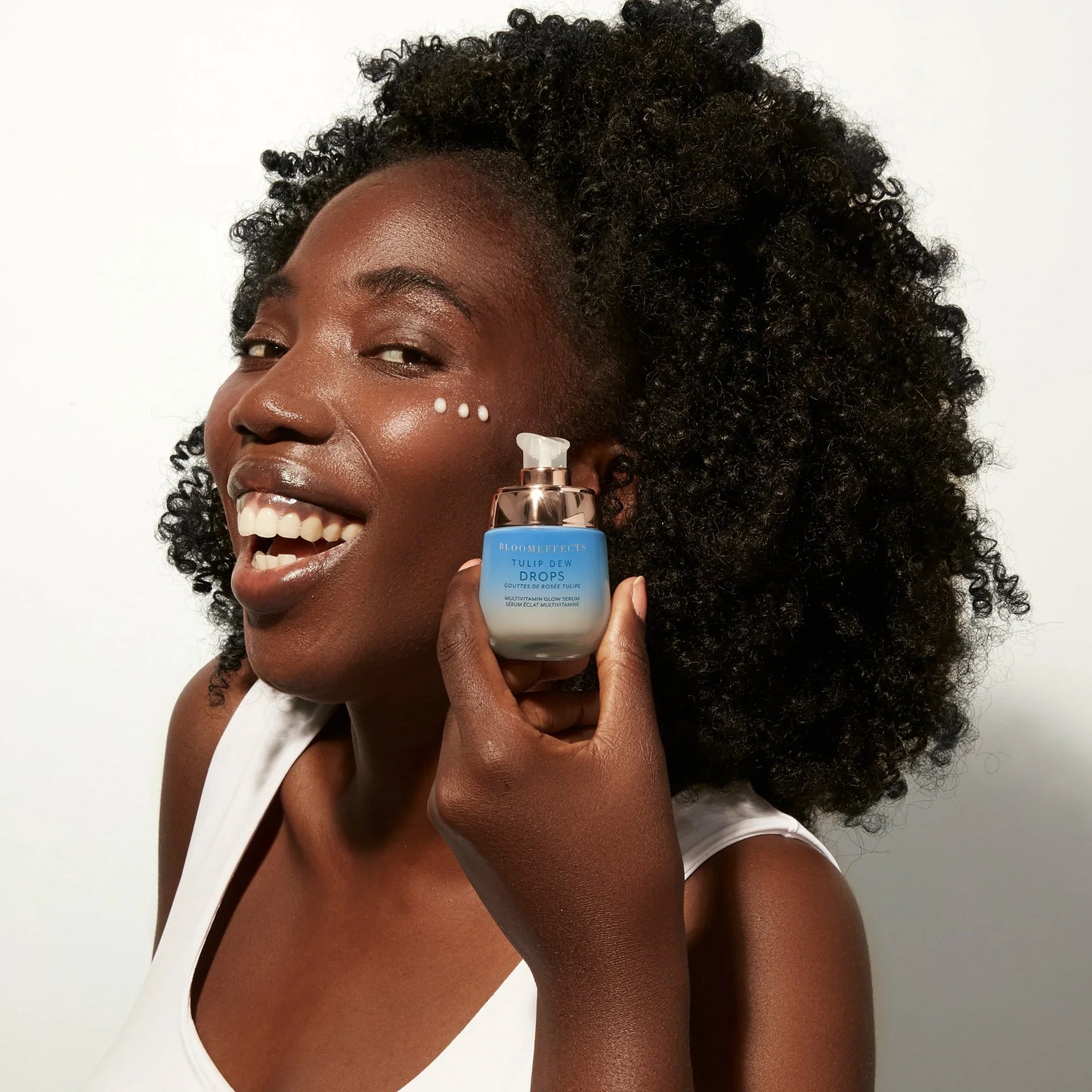 Model with black curly hair is holding a bottle with pump next to her face. She is holding BLOOMEFFECTS Tulip Dew Drops