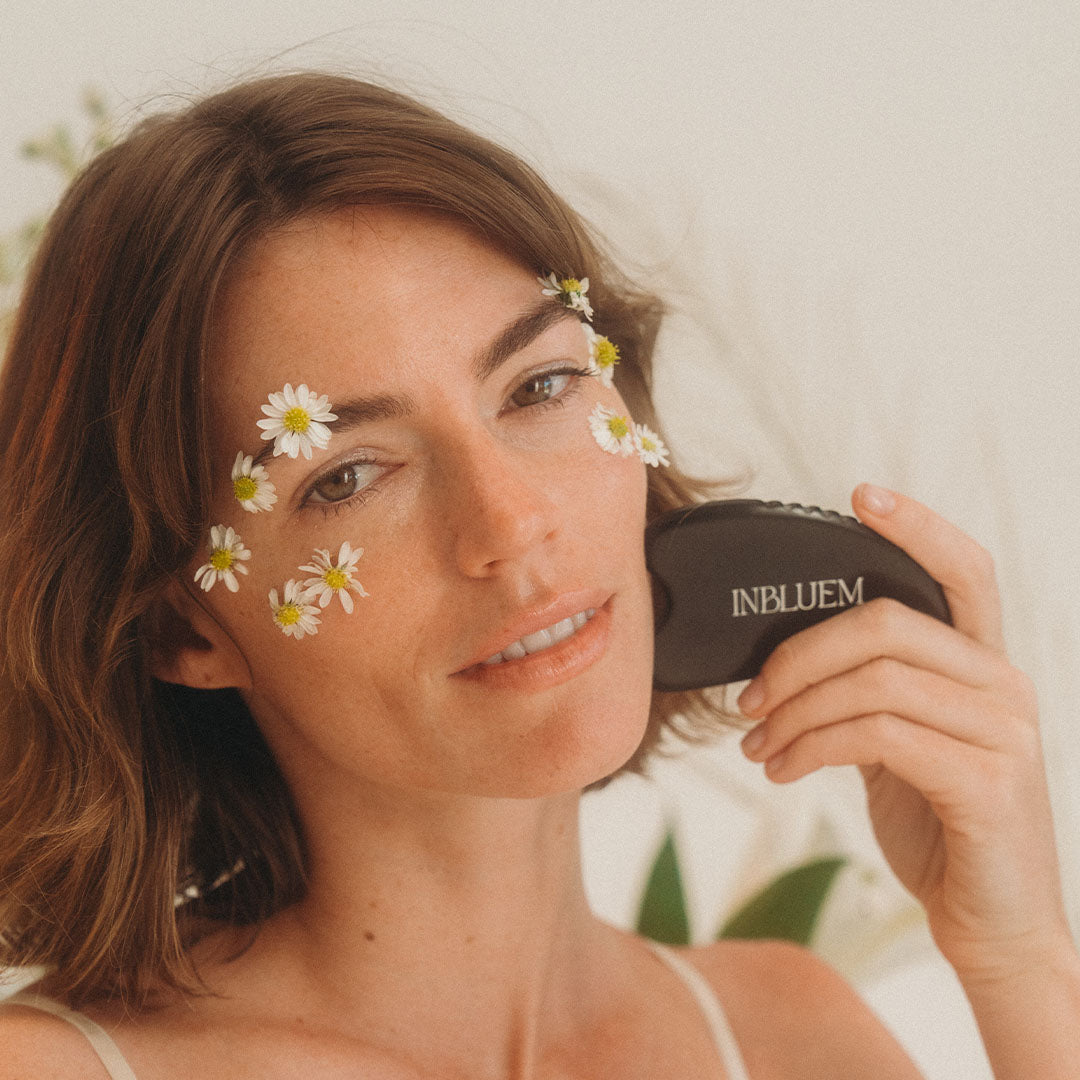 Woman with flowers around her eyes is holding an INBLUEM gua sha against a blurred background. INBLUEM Bian Stone Gua Sha