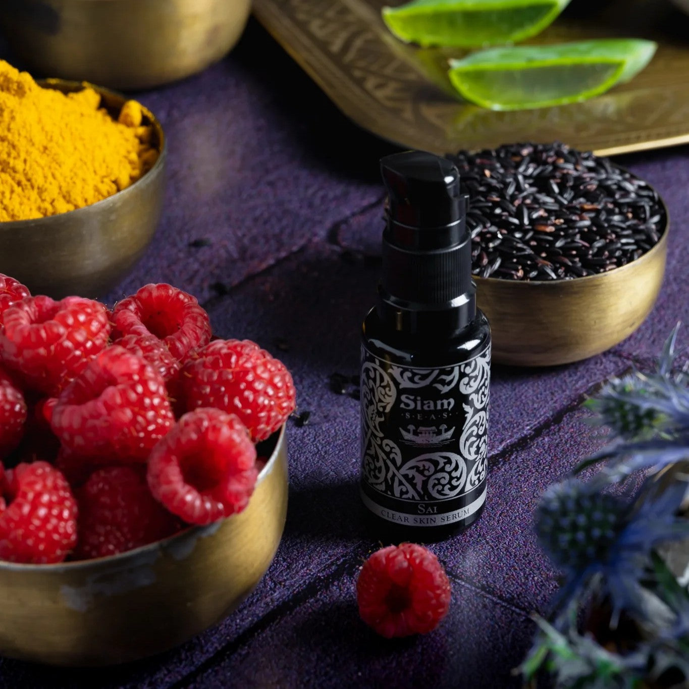 Bottle with pump of Siam Seas Sai Clear Skin Vitamin C Serum with bowls of raspberries, black rice, and yellow powder on a purple surface.