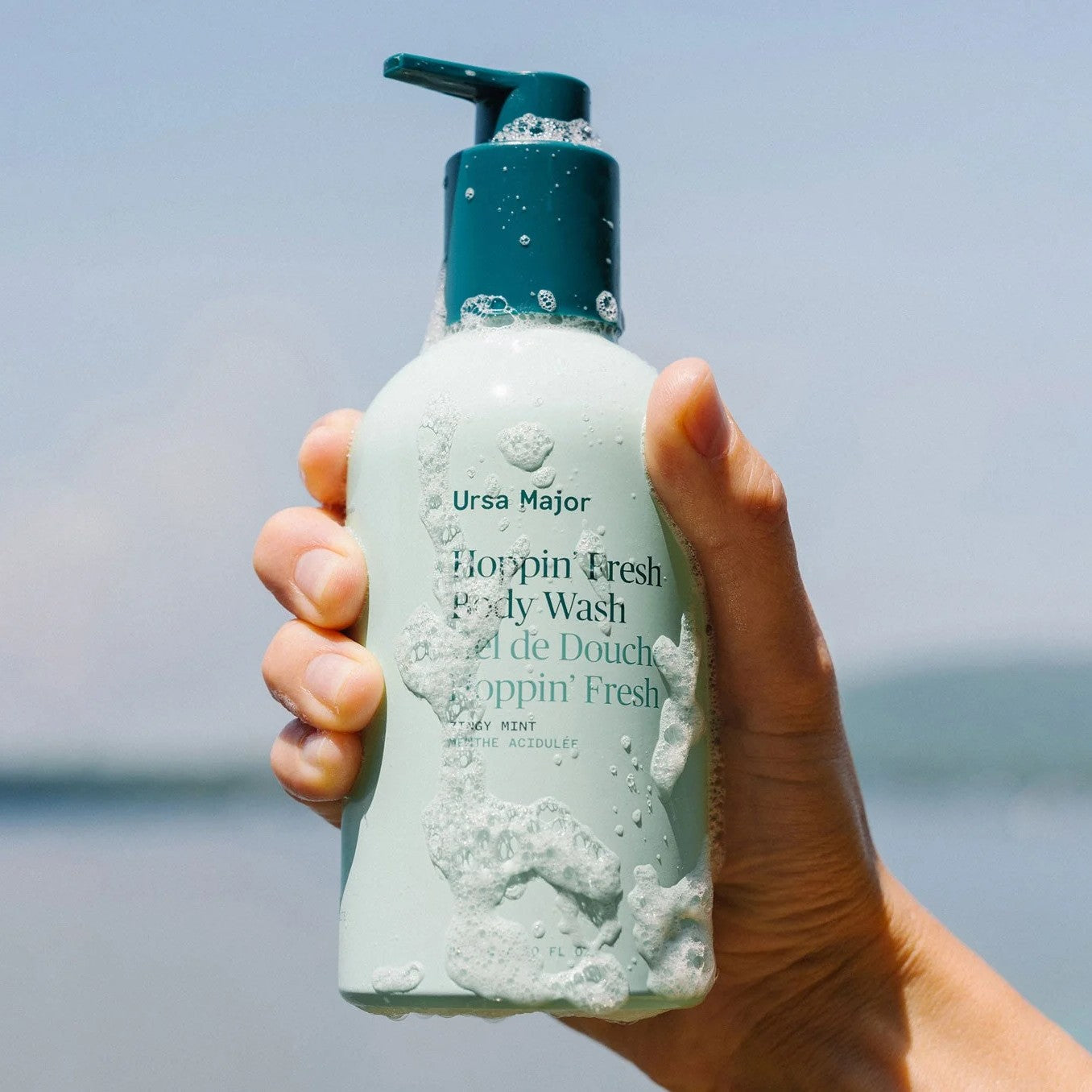 Hand holding a bottle of Ursa Major body wash with a blurred natural background
