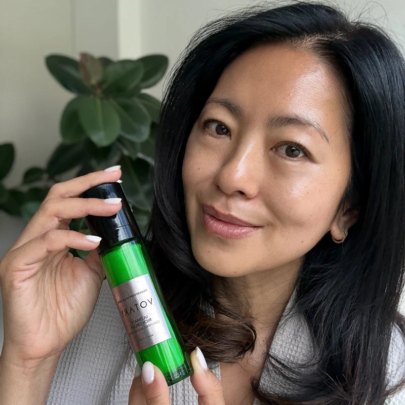 Woman holding a green skincare product with a plant in the background. VOTARY Ultimate Rich Cream