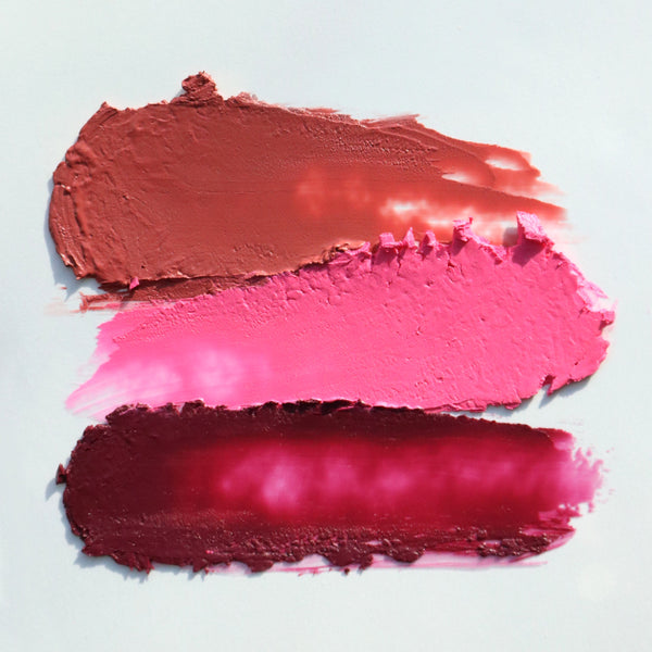 Three horizontal lipstick swatches showing different shades of pink and red cream makeup on a light gray background.