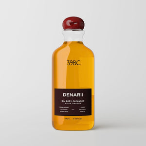 A clear bottle of 39BC Denarii oil body cleanser filled with golden yellow liquid, featuring a dark brown label and a glossy dark red cap, set against a light gray background.