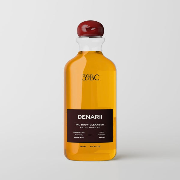 A clear bottle of 39BC Denarii oil body cleanser filled with golden yellow liquid, featuring a dark brown label and a glossy dark red cap, set against a light gray background.