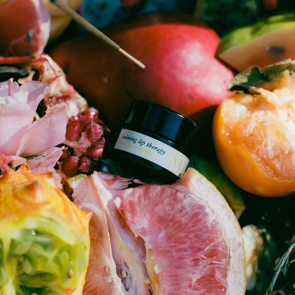 Close-up of a natural skincare balm in a small jar with ingredients including vanilla bean, lavender, and cacao. The balm is surrounded by vanilla beans, lavender sprigs, and cacao nibs, emphasizing its natural and nourishing components.