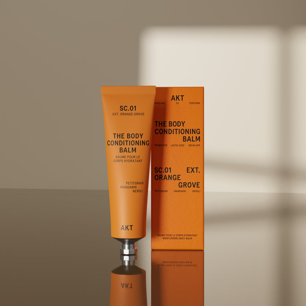 A squeeze tube of AKT London The Body Conditioning Balm in scent 01 ORANGE GROOVE, featuring a vibrant orange body and a metallic silver cap, set against a clean off-white background.