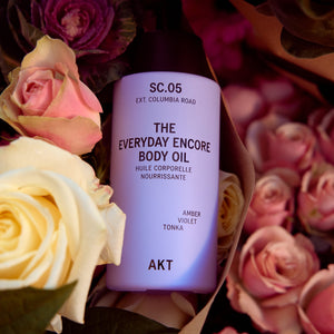 A bottle of AKT The Everyday Encore Body Oil in scent SC.05 Columbia Road, styled in a flat-lay arrangement with pink and cream roses.