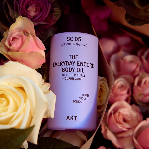 A bottle of AKT The Everyday Encore Body Oil in scent SC.05 Columbia Road, styled in a flat-lay arrangement with pink and cream roses.