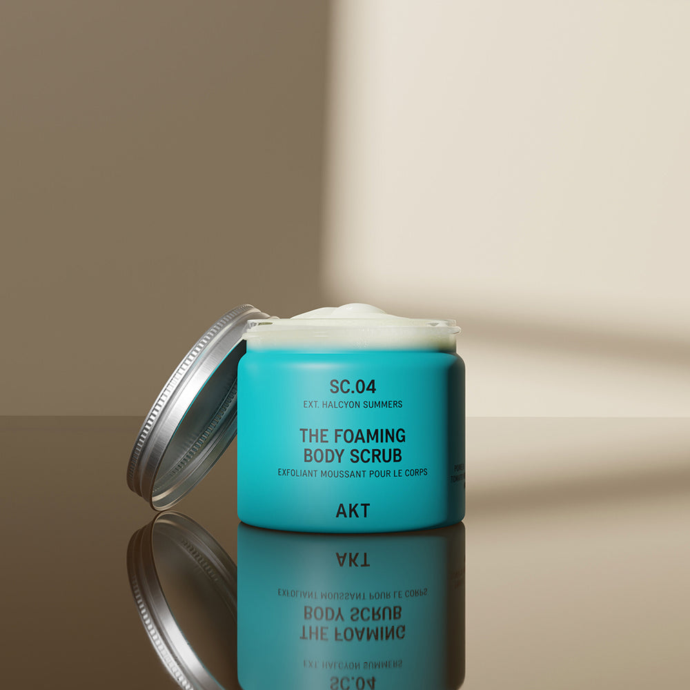 A turquoise blue jar of AKT London The Foaming Body Scrub with a silver metal lid, on a solid white background.