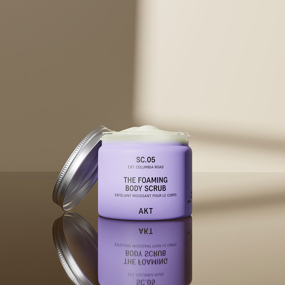 A lavender purple jar of AKT London The Foaming Body Scrub with a silver metal lid, set against a clean off-white background with a soft shadow.