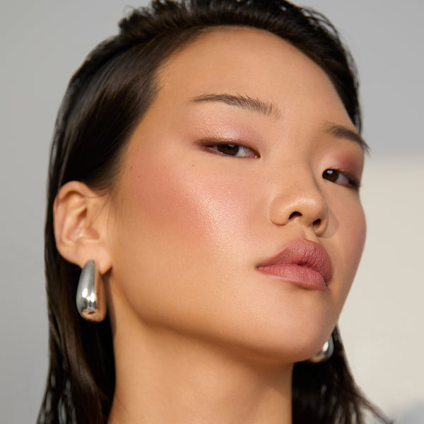 A close-up portrait of a model wearing a large, polished silver teardrop-shaped earring on her left ear, with her face filling the frame against a soft, neutral background.