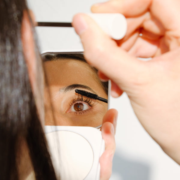 Person applying ALEPH Lift / Lengthen Mascara to their eyelashes in front of a mirror.