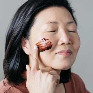 Woman applying a face mask on her cheek, with a neutral background. APOTERRA Hibiscus Exfoliating Mud