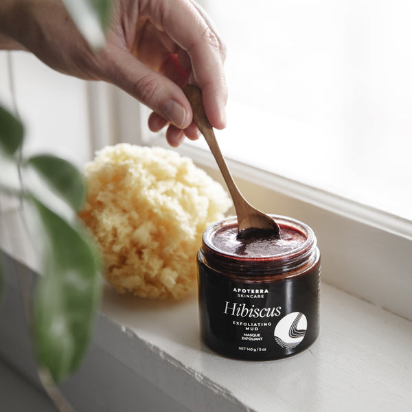 Hand scooping a jar of Hibiscus exfoliating mud by Apoterra with a wooden spoon, placed on a windowsill.