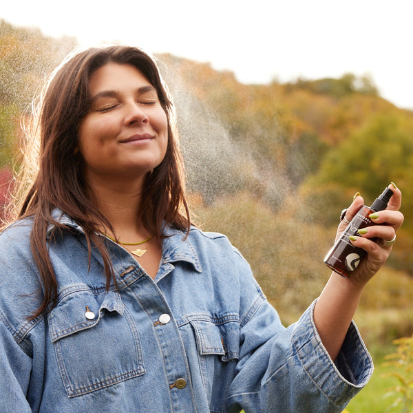 Woman in a denim jacket holding a spray bottle outdoors with trees in the background. APOTERRA Neroli Clarifying Complexion Mist