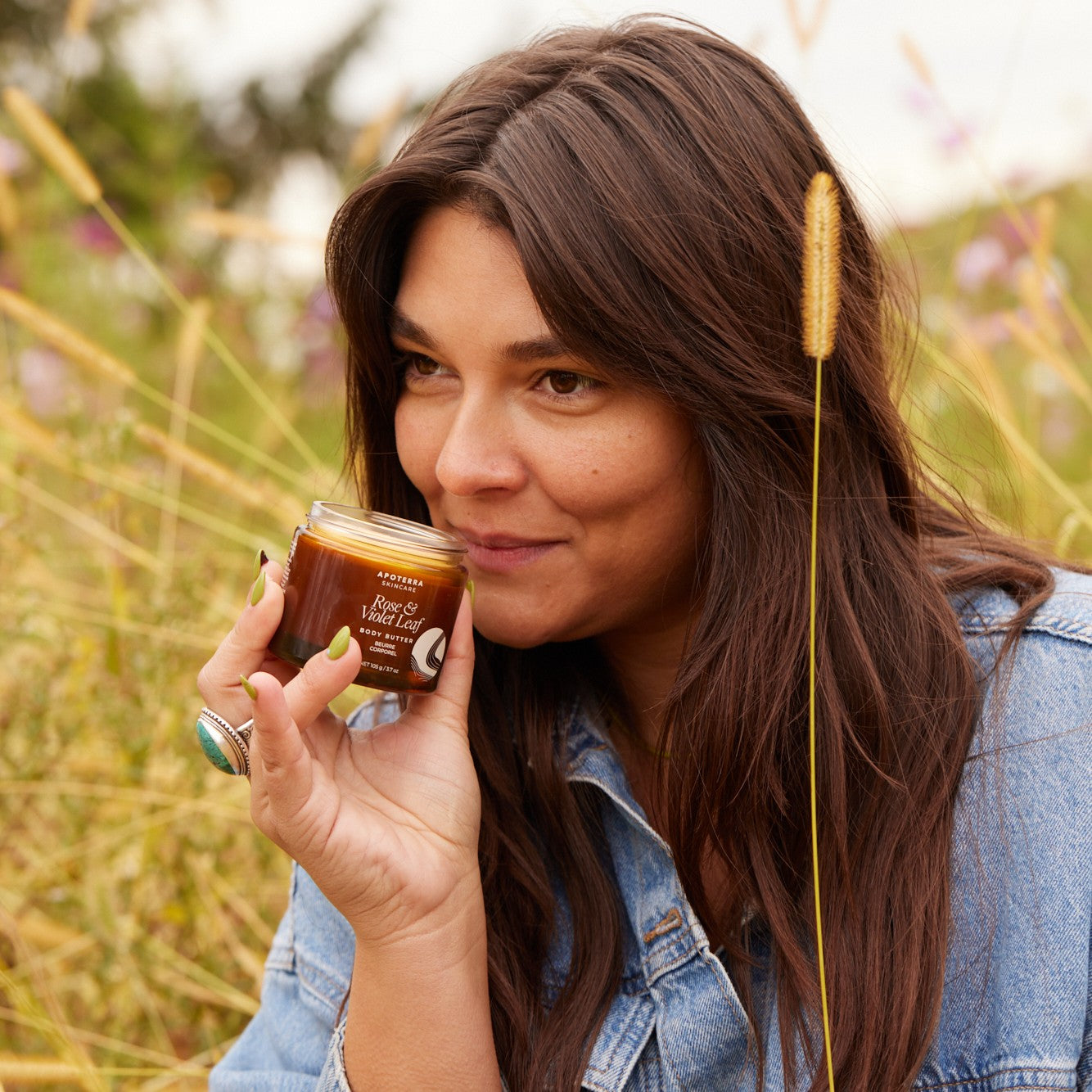 Woman holding a jar of APOTERRA Rose & Violet Leaf Body Butter in a field with tall grass and wildflowers.
