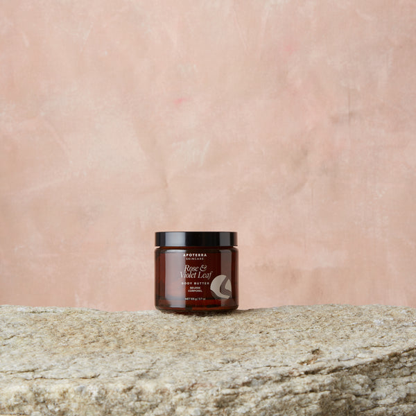 Jar of body butter on a stone surface with a pink textured background. Apoterra Rose & Violet Leaf Body Butter
