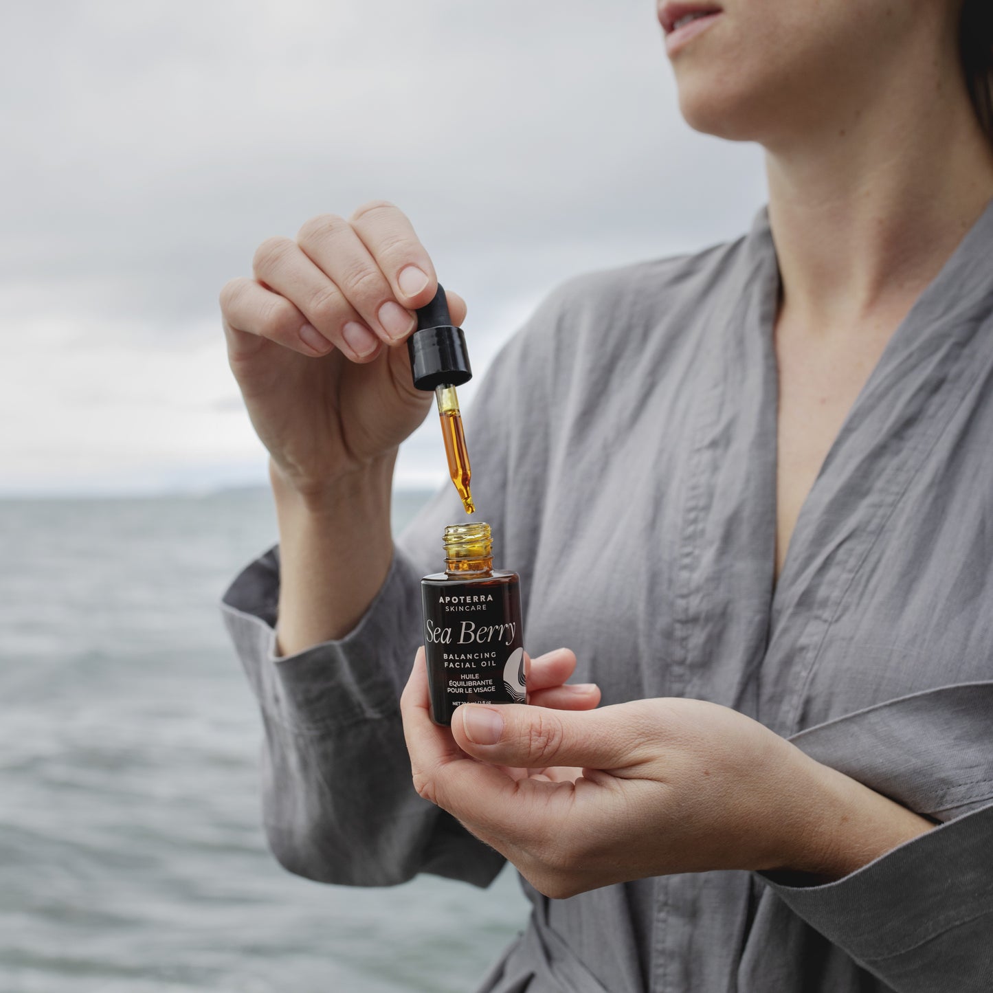 Woman holding a bottle of Sea Berry oil with a dropper over water.  APOTERRA Sea Berry Balancing Facial Oil
