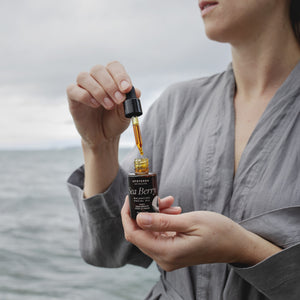 Woman holding a bottle of Sea Berry oil with a dropper over water.  APOTERRA Sea Berry Balancing Facial Oil