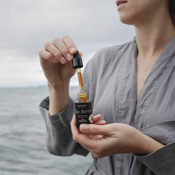 Woman holding a bottle of Sea Berry oil with a dropper over water.  APOTERRA Sea Berry Balancing Facial Oil