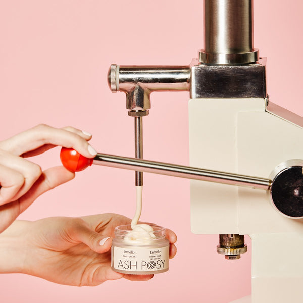 Hand holding a small container of cream labeled ASH POSY Lumello Face Creme under a faucet with pink background