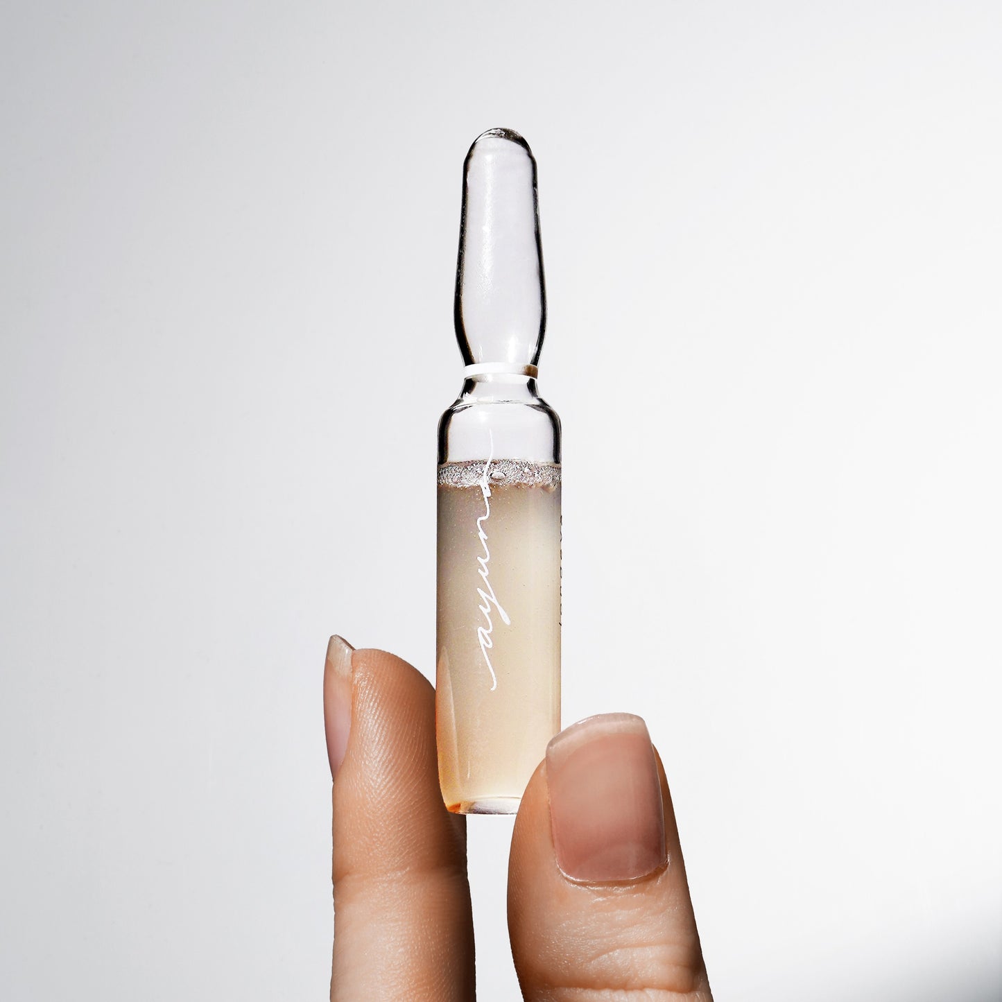 Hand holding a skincare ampoule with a clear liquid on a light gray background