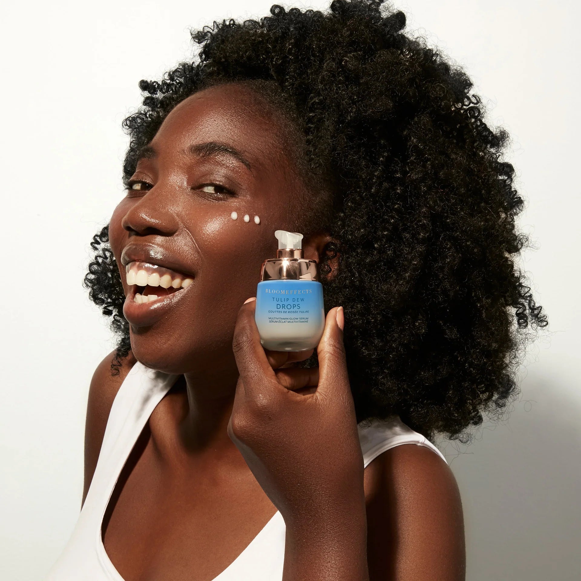 Model with black curly hair is holding a bottle with pump next to her face. She is holding BLOOMEFFECTS Tulip Dew Drops