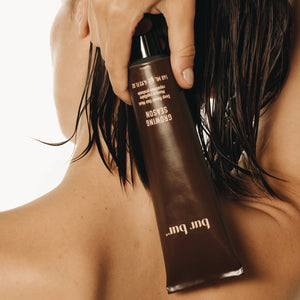 Woman holding a brown bottle labeled Bur Bur hair mask at the back of her neck/short bob dark hair against a neutral background