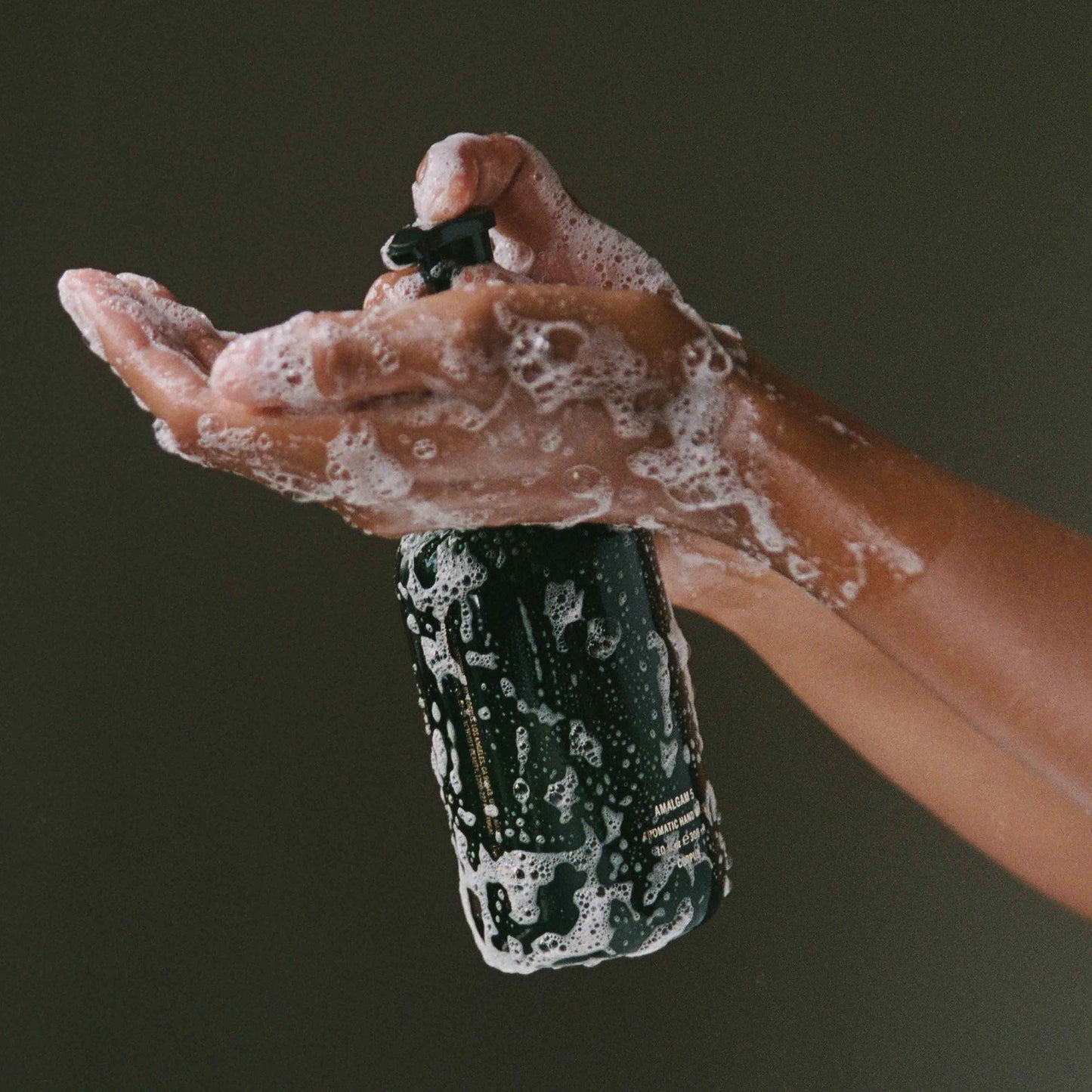 Hand holding a black bottle with soapy hands against a dark background. CORPUS Aromatic Hand Wash Amalgam 7