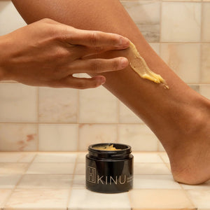 Person applying H IS FOR LOVE KINU Glowing Body Balm to leg with a jar of balm in the foreground, tiled bathroom background