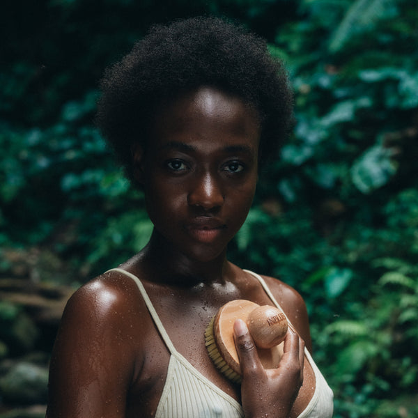 Woman holding a wooden brush against a green leafy background. INBLUEM Lotus Wood Body Brush