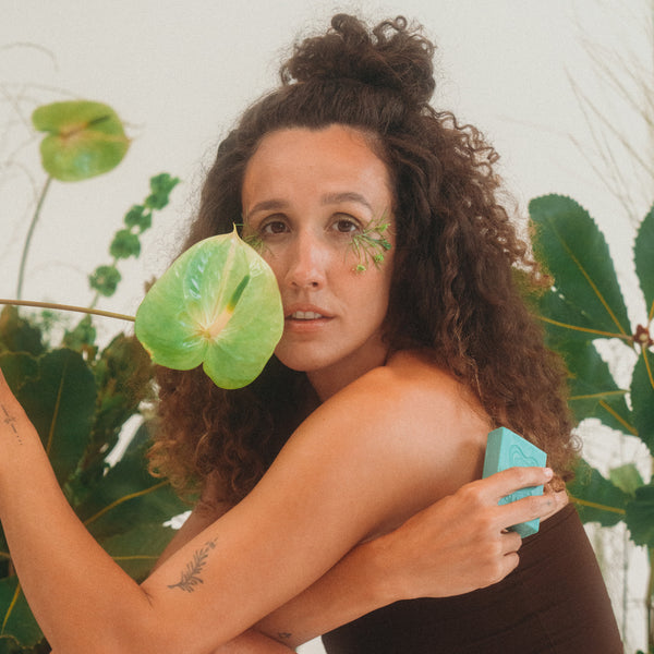 Woman with green leaves on her face and shoulders, surrounded by plants. she is holding INBLUEM Soul Soap Finger Lime & Eucalyptus in her hand.