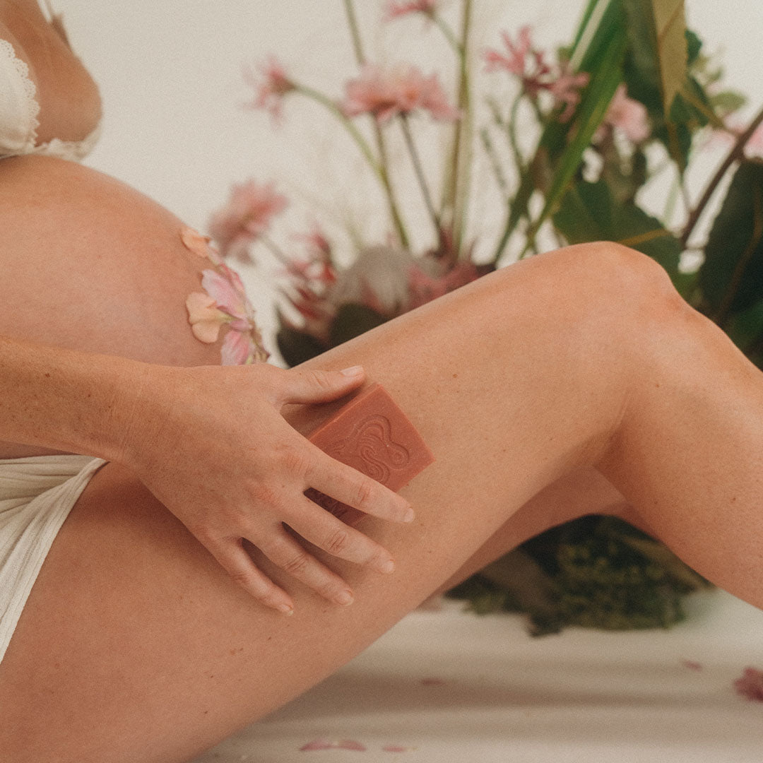 A pregnant woman is applying a red bar of soap to her leg with a blurred floral background. INBLUEM Soul Soap Kakadu Plum & Kunzea