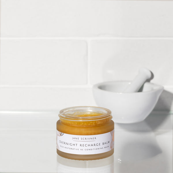 A small glass jar of Jane Scrivner Overnight Recharge Balm sits on a white countertop with a white mortar and pestle blurred in the background against a white tiled wall.