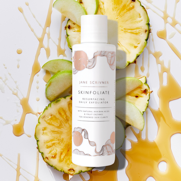 A bottle of Jane Scrivner Skinfoliate Resurfacing Daily Exfoliator centered on a white background, surrounded by sliced pineapple and green apple pieces with caramel-colored syrup drizzle.