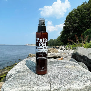 Bottle of 'Patio Oil Sunset Blend' on a rock by a body of water with trees in the background.
