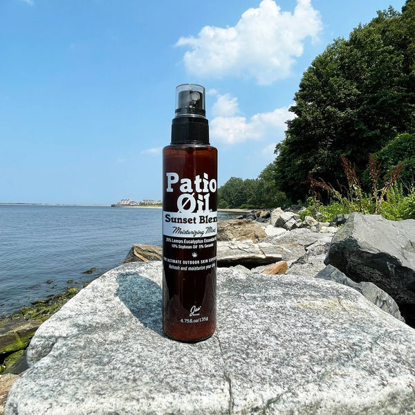 Bottle of 'Patio Oil Sunset Blend' on a rock by a body of water with trees in the background.