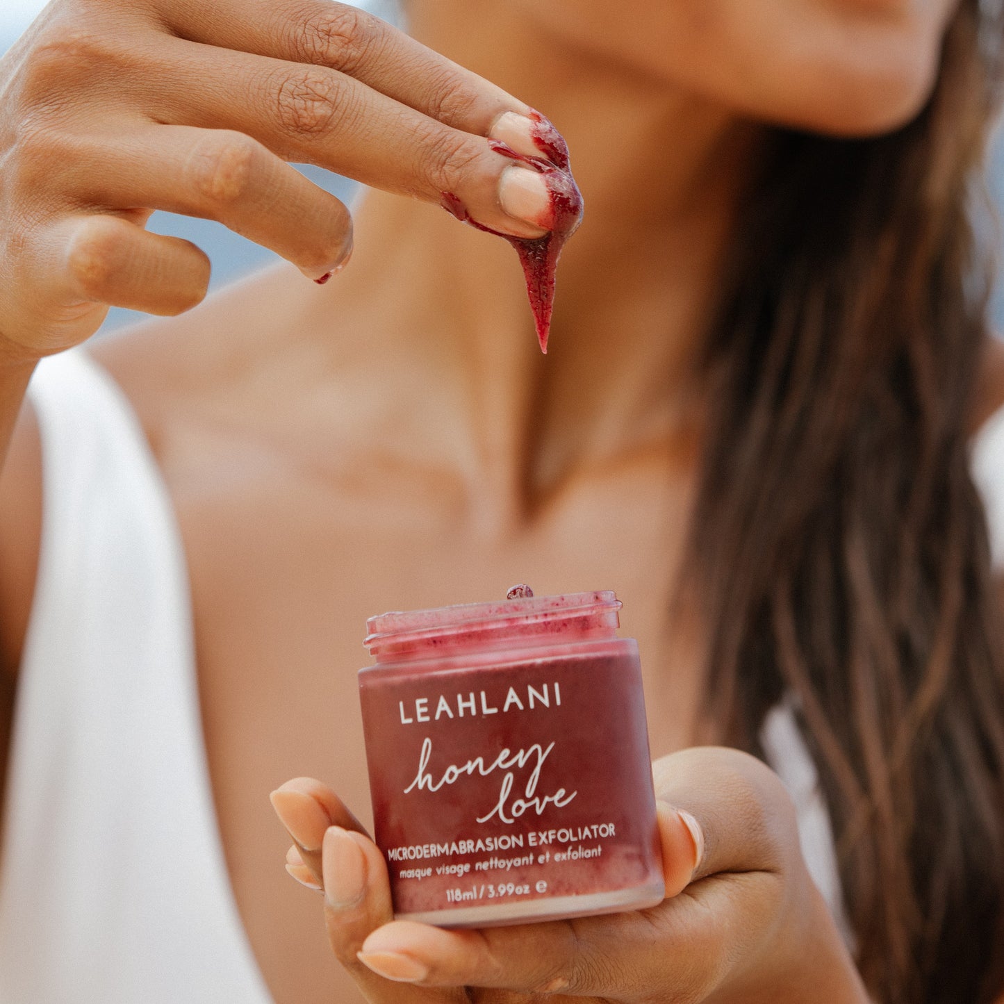 Person applying a red exfoliant to their skin with a jar of Leahlani honey love exfoliant in the foreground. LEAHLANI SKINCARE Honey Love Exfoliator ALWAYS SHOW