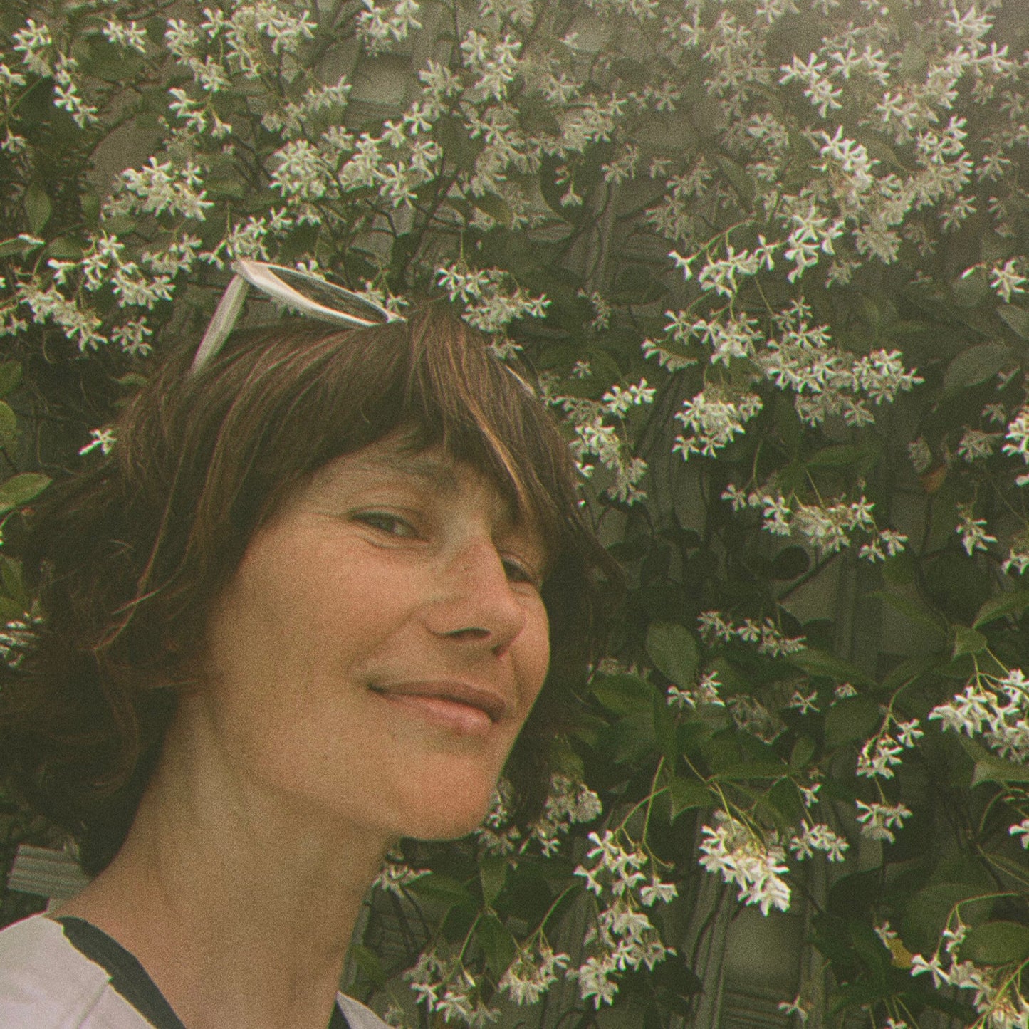 A woman with sunglasses on head standing in front of a flowering bush of jasmine