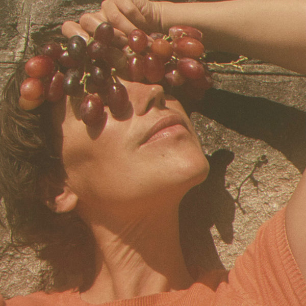 A woman holding red grapes and placing them on her forehead against a textured wall. LEPAAR Summer Wine / Even Tone Antioxidant Shield / Zero Essential Oils ALWAYS SHOW