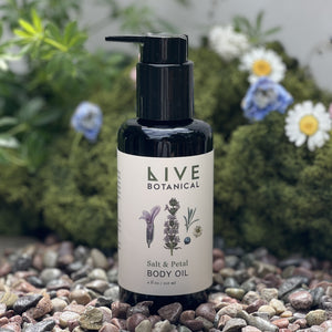 Bottle of Live Botanical Salt & Petal Body Oil on a pebble surface with plants and flowers in the background