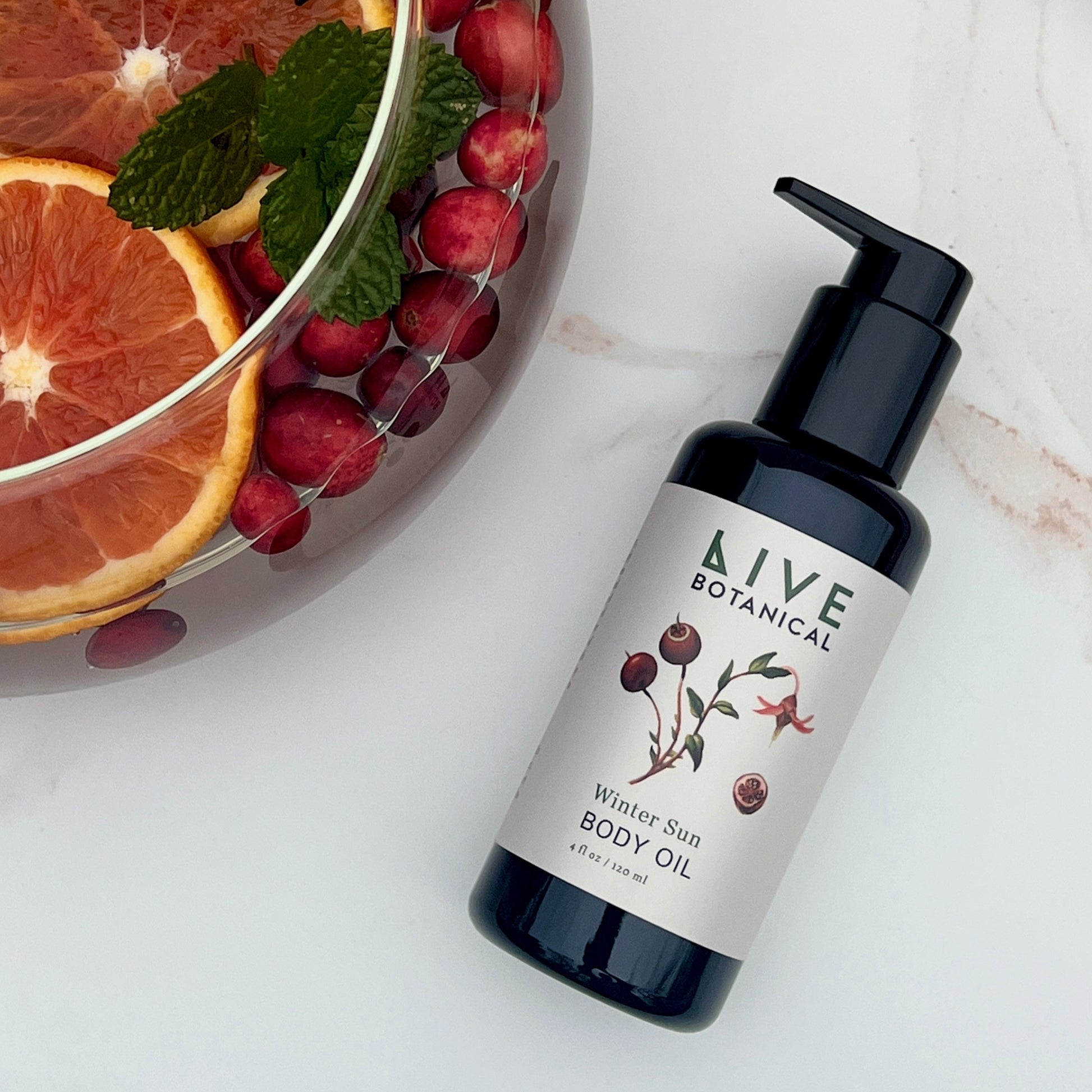 Bottle of Live Botanical body oil with oranges and cranberries on a marble surface