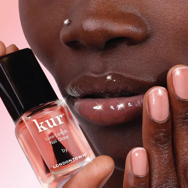 Nail polish bottle of LONDONTOWN Luxe Length Nail Gloss held by a hand with pink nail polish against a pink background