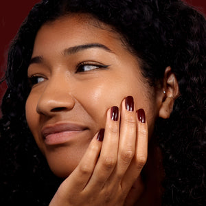 Woman with dark curly hair and dark red nail polish touching her face against a dark background