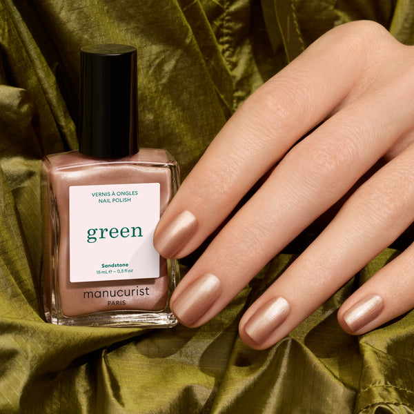 A hand with manicured nails painted in a metallic champagne pink polish rests beside a bottle of Manucurist nail polish in color Sandstone on draped olive green fabric.