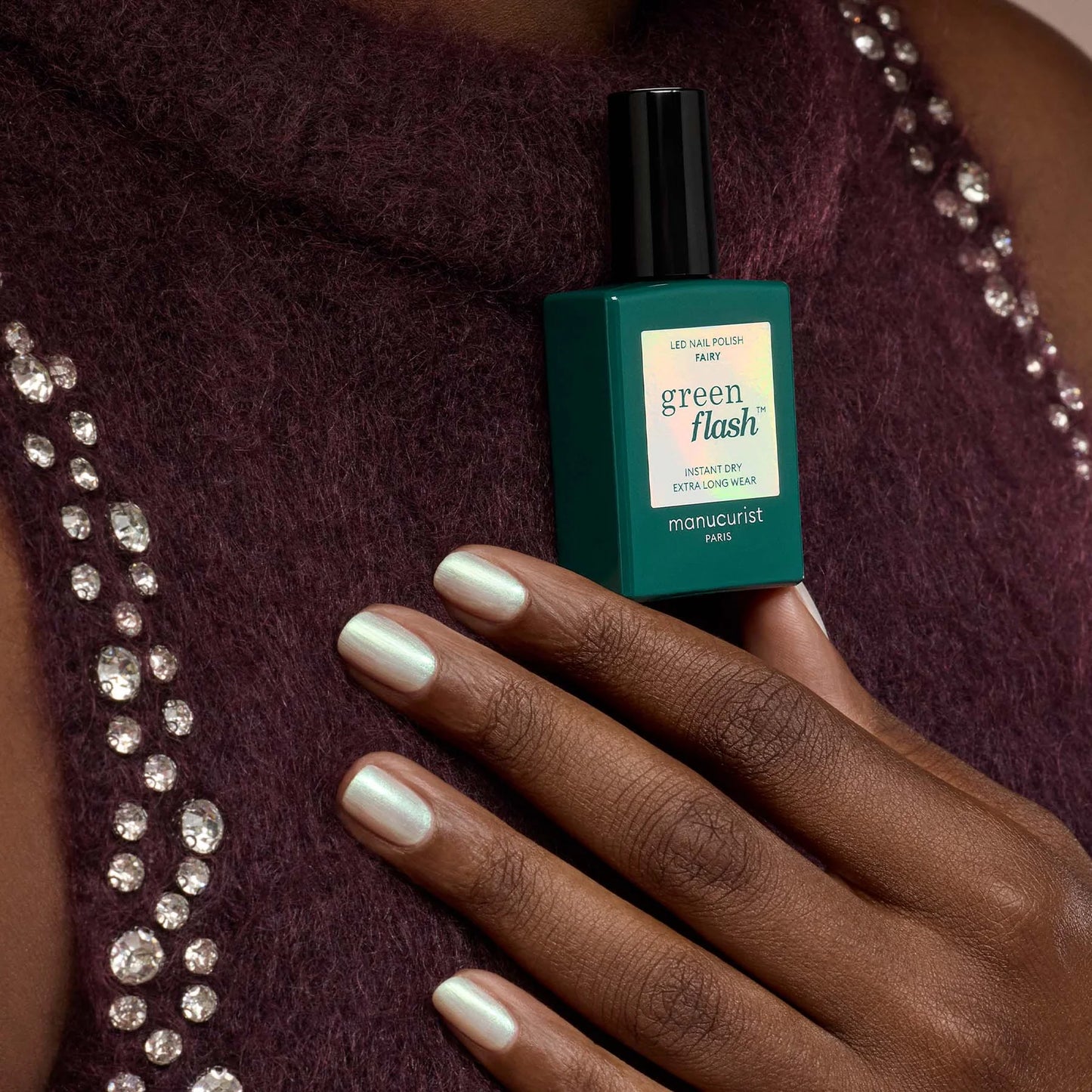 Green Flash nail polish bottle held by a hand against a textured fabric background