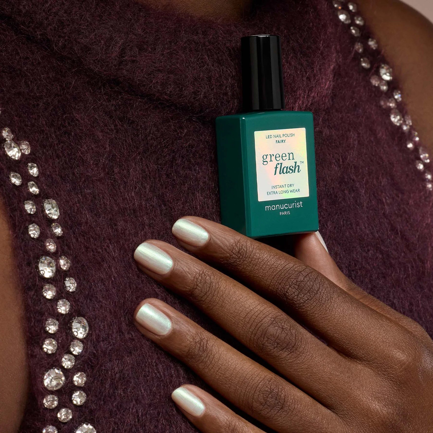 Green Flash nail polish bottle held by a hand against a textured fabric background