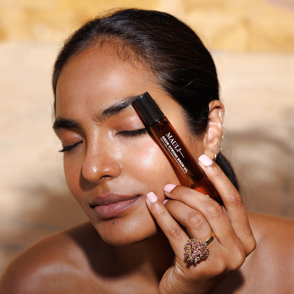 Woman applying MAULI RITUALS Grow Strong Brow Oil to her face with a blurred background